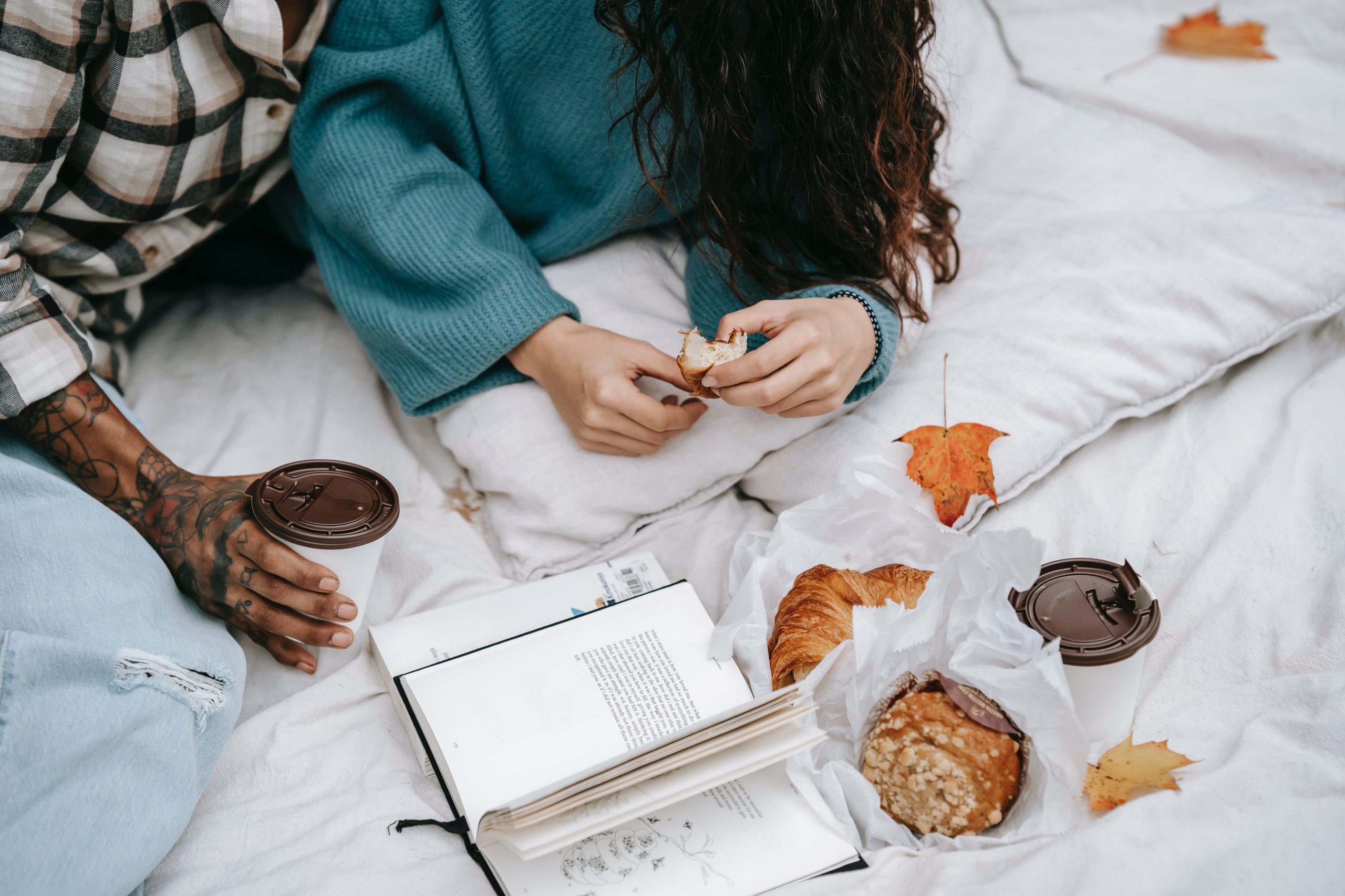 a couple sitting on a blanket on the ground reading, drinking coffee and snacking on croissants